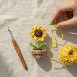 May include: A handmade crochet sunflower in a small pot, with yellow petals and a brown centre. A crochet hook and a ball of yellow yarn are also visible. The sunflower is approximately 7.6 cm tall.