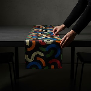 Op de afbeelding: Een zwarte tafelloper met een kleurrijk, abstract ontwerp van gebogen vormen in blauw, oranje, groen en wit. De loper wordt op een donkere tafel geplaatst. Het thema is woondecoratie.