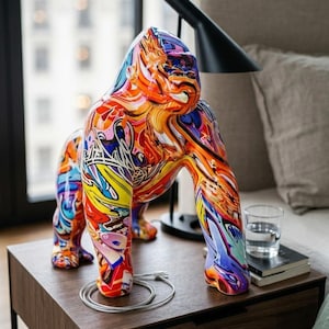 Puede incluir: Una colorida escultura de gorila con un diseño abstracto vibrante en tonos de rojo, naranja, amarillo, azul y morado. La escultura está sobre una mesita de noche de madera junto a una lámpara y un vaso de agua. El gorila mide unos 30 cm de altura.