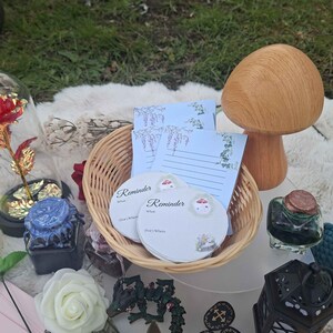 May include: A collection of whimsical items, including a wooden mushroom, a woven basket with notepads, and decorative objects. The notepads have the word "Reminder" on them. Other items include a rose, a small lantern, and a glass bottle.
