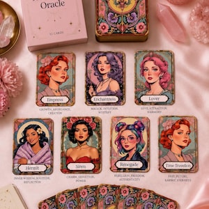 May include: A pink box set of "The Archetype Oracle" cards with 12 cards. The cards feature illustrations of women with titles like "Empress" and "Enchantress". The card backs and box have floral designs. A lit candle and crystals are also visible.