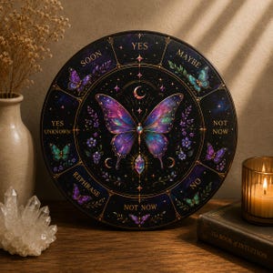 May include: A round, black divination board with a large, colorful butterfly in the center. The board features text such as "Yes," "Maybe," and "Not Now," along with crescent moons and stars. The board is surrounded by floral and butterfly illustrations.