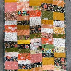 May include: A patchwork quilt with a variety of floral and geometric patterns in shades of pink, orange, yellow, green, and black. The quilt is bordered with a dark green floral fabric and has a quilted texture.