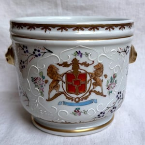Peut inclure: Un cache-pot en porcelaine blanche orné d'un blason central avec deux lions entourant un écu rouge. Le récipient présente des volutes blanches en relief, des motifs floraux peints à la main en violet, rose et vert, et un liseré doré.