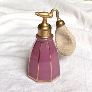 May include: A vintage perfume atomizer featuring a faceted, mauve-colored glass bottle with gold-toned metal trim. The top assembly includes a gold-toned metal nozzle and a textured, off-white fabric bulb attached by a curved metal arm.