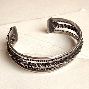 May include: Silver cuff bracelet featuring a layered design. The bracelet has multiple bands with a combination of smooth and textured elements, including a rope-like pattern. The open design allows for easy wear.