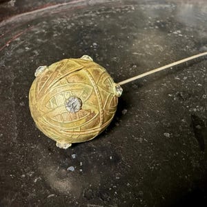 May include: A gold-toned spherical hat pin featuring an intricate etched leaf pattern across its surface. The round head is accented with small, clear, faceted crystals embedded at intervals, and it is attached to a long, thin, straight metal pin.