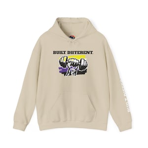 May include: A beige hoodie with a front pocket and a drawstring hood. The front features the text "BUILT DIFFERENT." above a graphic of a weight being lifted by a fist. The right sleeve has the text "BUILT DIFFERENT." printed vertically.