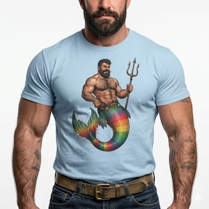 May include: Light blue t-shirt featuring a muscular merman illustration. The merman has a beard, holds a trident, and has a rainbow-colored tail. The shirt has a crew neck and short sleeves.
