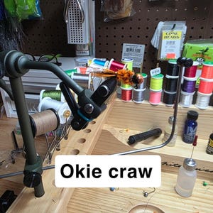 May include: A fly tying setup with an "Okie craw" fly in progress. The fly is orange and brown, mounted on a black vise. Spools of thread in various colors and other fly tying materials are visible.