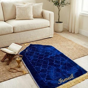May include: A blue prayer rug with a geometric pattern and gold fringe. The word "Ismail" is embroidered in gold. The rug is placed on a light wood floor, near a sofa and a small table with a book.