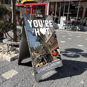 Può includere: Un cartello a forma di A con la scritta "YOU'RE HOT" in bianco, con un simbolo a forma di cuore al posto della "O". Il cartello riflette una scena di strada con edifici e persone. La parte inferiore del cartello recita "@THEFULLROUTINE".