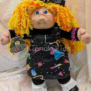 May include: A Cabbage Patch Kids doll with yellow curly hair, glasses, and a black dress with colorful geometric shapes. The doll has a black bow, black shoes, and white lace socks. The tag reads "Cabbage Patch Kids". The text "Kipnuk Kids" is also visible.