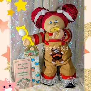 May include: A Cabbage Patch Kids doll with red yarn pigtails, wearing a brown and red outfit with a teddy bear applique. The doll holds a yellow toy and a pacifier. Includes a birth certificate and adoption papers.