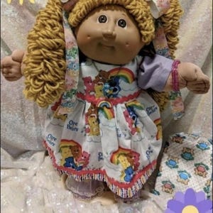 May include: A vintage Cabbage Patch Kids doll with blonde yarn pigtails and a painted face. The doll wears a white dress printed with Care Bears and rainbows. The dress has a red trim and the text "Care Bears".