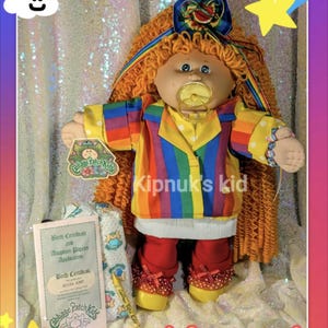 May include: A Cabbage Patch Kids doll, Julia Amy, with orange yarn hair, a rainbow striped shirt, red pants, and yellow shoes. The doll has a pacifier and a birth certificate. The text "Kipnuk's Kid" is displayed on the image.