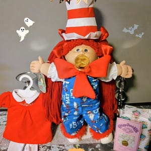 May include: A Cabbage Patch Kids doll dressed in a Cat in the Hat costume, complete with a red and white striped hat, red bow tie, and blue pajamas. A red outfit and a package of diapers are also displayed. Halloween decorations are in the background.