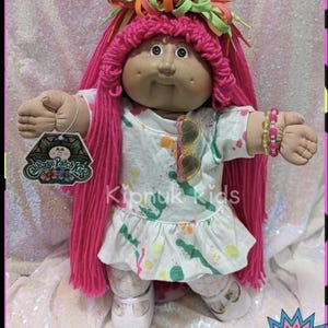 May include: A Cabbage Patch Kids doll with pink yarn hair and a colorful headpiece. The doll wears a white dress with a colorful splatter design and sunglasses. The doll has a tag that says "Cabbage Patch Kids" and the text "Kipnuk kids".