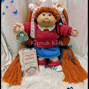 May include: A Cabbage Patch Kids doll with red yarn hair, wearing a red sweater, blue skirt, and white shoes. The doll has a birth certificate and a tag. The image includes the text "Kipnuk kids".