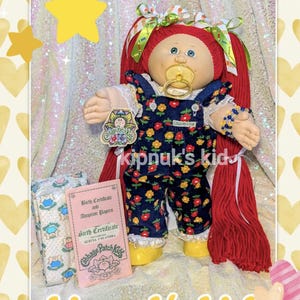 May include: A Cabbage Patch Kids doll with red yarn hair, wearing a navy blue floral jumpsuit and yellow shoes. The doll has a pacifier and a birth certificate. The text "kipnuk's kid" is visible.