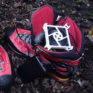 May include: A pair of red and black hiking boots with white plastic fan inserts placed inside the openings. The fans are connected by black wires to a small black rectangular power bank resting on the side of one of the boots.