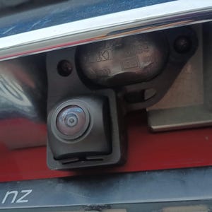 Może przedstawiać: Zbliżenie kamery cofania samochodu, zamontowanej na czarnym wsporniku. Obiektyw kamery jest widoczny, otoczony ciemną obudową. Tło obejmuje czerwoną powierzchnię i chromowane wykończenie.