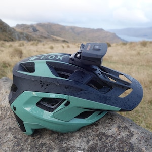 May include: A matte black and sage green bicycle helmet with the word FOX printed on the side. The helmet features multiple ventilation cutouts and a black action camera mounted securely to the top centre of the shell, positioned above the visor.