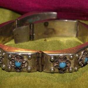 May include: A silver bracelet with a hinged clasp and decorative details. The bracelet is adorned with turquoise-colored stones set in silver.