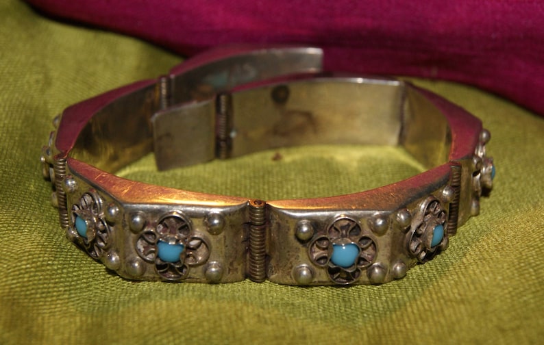 May include: A silver bracelet with a hinged clasp and decorative details. The bracelet is adorned with turquoise-colored stones set in silver.