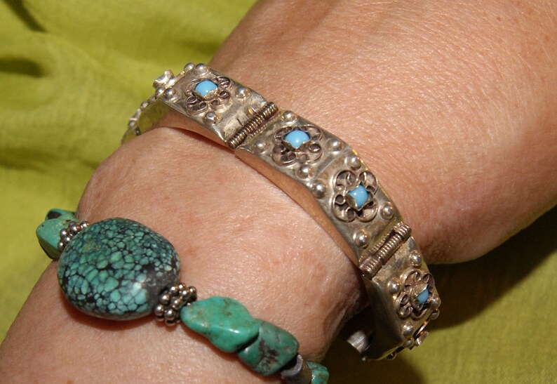 May include: A silver bracelet with turquoise stones and a silver and turquoise beaded bracelet on a person's wrist.