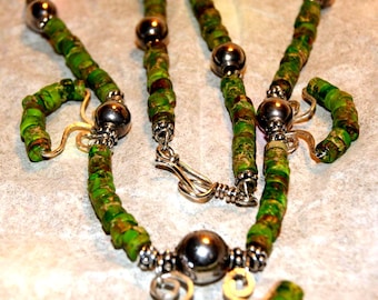 May include: A handmade necklace and matching earrings set. The jewelry features green and brown beads with silver-colored accents and a clasp. The earrings have a hook closure. The necklace has a large, round bead in the center.