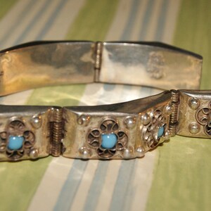 May include: A silver bracelet with a hinged clasp and decorative filigree. The bracelet is adorned with turquoise blue stones.
