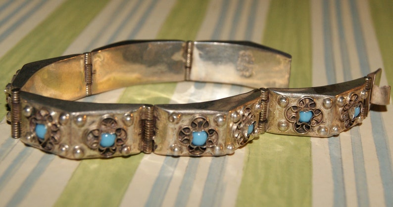 May include: A silver bracelet with a hinged clasp and decorative filigree. The bracelet is adorned with turquoise blue stones.