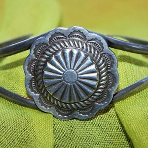 Puede incluir: Un brazalete de plata con un diseño circular decorativo. El diseño presenta un patrón de rayos de sol con un círculo más pequeño en el centro. El brazalete está hecho de dos bandas de metal.