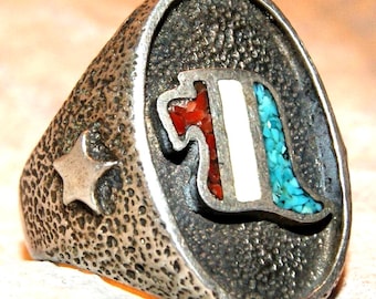 May include: A silver-toned ring with a textured, pitted surface featuring a raised star on the side. The oval face displays an inlaid design with three vertical sections containing crushed red stone, a smooth white strip, and crushed blue turquoise.