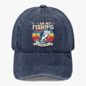 May include: A navy blue baseball cap with a curved brim. The cap features a colorful graphic of a fish jumping out of water with the text "I AM NOT FISHING I AM THINKING ABOUT IT".