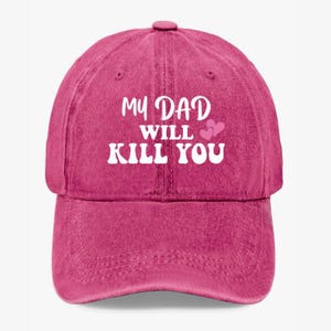 May include: A magenta baseball cap with the text "MY DAD WILL KILL YOU" in white, along with two pink hearts. The cap has a curved brim and a fabric strap closure.