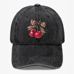 May include: A black denim baseball cap featuring a vibrant graphic of two red cherries tied with a leopard print bow. The cap has a curved brim and an adjustable strap for a comfortable fit. This hat is a stylish accessory.