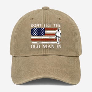 May include: A tan baseball cap featuring the American flag and a soldier graphic. The text "DON'T LET THE OLD MAN IN" is printed above and below the flag design. The cap has a curved brim and a button on top.