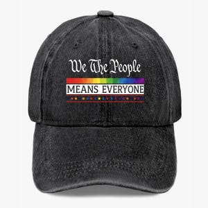May include: A black denim baseball cap with the text "We The People MEANS EVERYONE" in white and rainbow colors. The cap has a curved brim and a button on top. The text is centered on the front of the cap.
