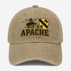 May include: A tan baseball cap featuring a graphic of an AH-64 Apache helicopter and a yellow shield emblem with a black horse silhouette. The text AH-64 APACHE is printed in bold black letters with yellow outlines across the front of the cap.