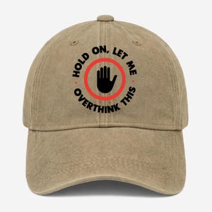 May include: A tan baseball cap featuring a black graphic of an open hand inside a red circle. The text "HOLD ON, LET ME OVERTHINK THIS" is printed in black capital letters in a circular arrangement around the central hand icon and red ring.