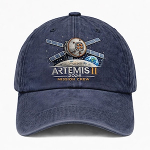 May include: A navy blue baseball cap featuring an Artemis II mission design. The design includes a spacecraft, Earth, the moon, and the text "ARTEMIS II 2026 MISSION CREW". The cap has a curved brim and an adjustable strap.