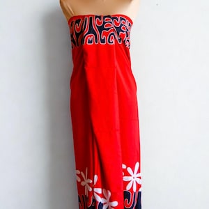 May include: A strapless sarong in a vibrant red color featuring a dark blue and white abstract pattern along the top edge and a dark blue and white floral motif along the bottom hem. The fabric is draped to create a long, flowing silhouette.