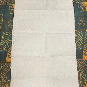 May include: A rectangular piece of handmade paper, folded into a long, narrow shape. The paper is off-white with visible texture and slight imperfections. The background features a patterned fabric with teal, gold, and brown colors.