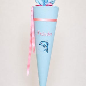 Puede incluir: Un cono escolar azul claro, con una cinta rosa y el nombre "Frieda" bordado. También presenta una ilustración de un delfín azul. La bolsa está atada con un lazo azul claro.