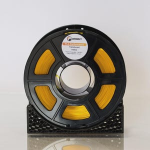May include: A black spool of yellow PLA Performance Translucent filament for 3D printing. The spool is labeled "ORGELY" and includes a sticker with product details. The filament is held by a black stand.