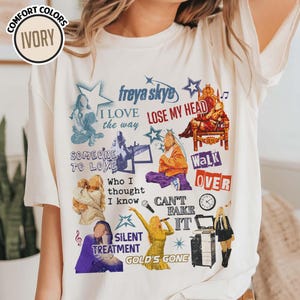May include: Ivory-colored t-shirt with a graphic design featuring the text "freya skye" and various song titles and illustrations in blue, orange, and gold. The shirt has a crew neck and short sleeves.
