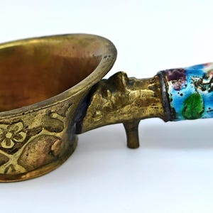 Op de afbeelding: Antieke messing schep met een bloemmotief op de kom en handvat. Het handvat is versierd met een blauw emaille bloemenpatroon. De schep heeft een kleine voet voor stabiliteit, waarschijnlijk een decoratief item. Afmetingen in cm.