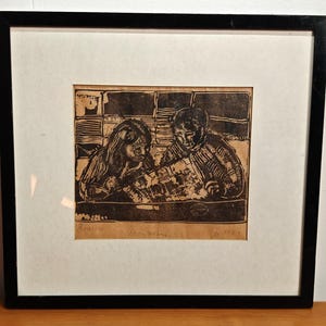 May include: A framed black and white woodcut print on tan paper depicting two figures seated at a table. The artwork is enclosed in a simple black frame with a white mat. The print features bold, textured lines and includes handwritten text at the bottom.